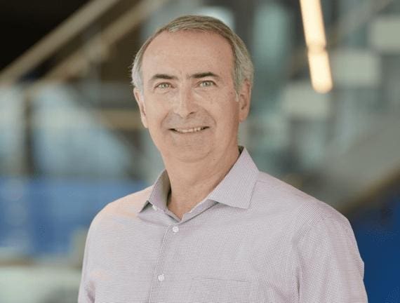 Smiling middle-aged man in a light purple shirt with blurred background.