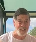 Smiling elderly man with gray hair and beard indoors near a window.