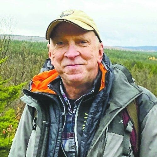 A man wearing outdoor gear and a cap, smiling in a natural setting.