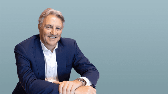 Smiling mature man in a suit sitting at a table against a blue background.